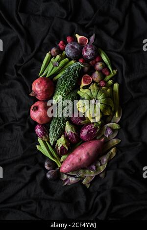 Top view of basket with assorted colorful fresh fruits and vegetables including pomegranate and plums and figs with mini eggplants and sweet potato on black background Stock Photo