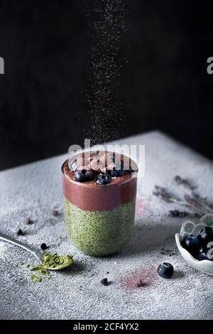 Glass of appetizing healthy matcha tea and chia seeds smoothie with chocolate and blueberries sprinkled with cocoa powder on dusted with sugar powder table Stock Photo