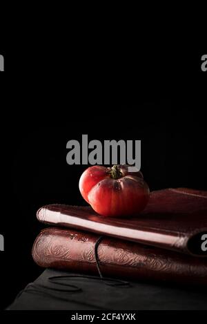 Ripe red tomato placed on stack of ancient books against black background Stock Photo