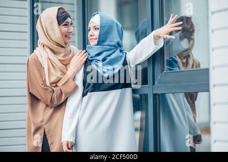 Two attractive women in hijab standing near the glass door. one girl routinely comes to side with your finger. outdoors Stock Photo