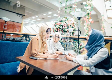 Three pretty muslim women with hijab in cafe. sitting on couches at a table and talking. One girl with glasses Stock Photo
