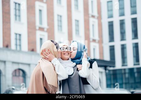 Two funny affectionate muslim women friends laughing and kissing girl in hijab an with glasses outdoors. Stock Photo