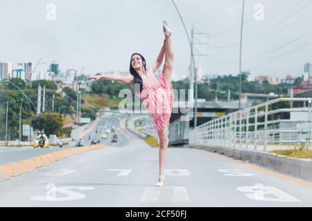 Full body young ballerina smiling and doing splits on empty lane of road while dancing in modern city outskirts Stock Photo