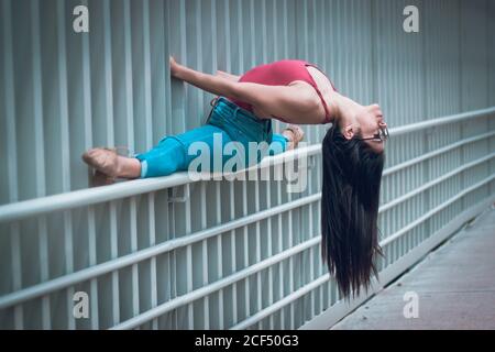 Side view of slip Woman in casual clothes and glasses doing splits on fence and bending back while dancing on street Stock Photo