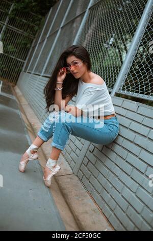 Side view of slim Woman ballerina in casual clothes and sunglasses sitting leaning on tiptoe with pointe shoes on brick wall fence on the street Stock Photo