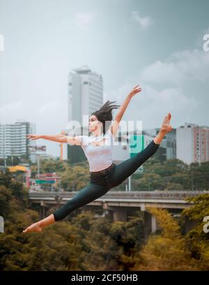Full body energetic barefoot Woman in casual clothes jumping and doing splits while dancing against contemporary city and cloudy sky Stock Photo
