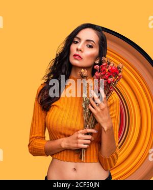 Confident young female model in trendy orange crop top holding bouquet of fresh flowers and looking at camera while standing against bright orange wall with geometric ornament in studio Stock Photo