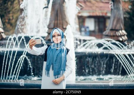 Young muslim woman in blue hijab making selfie with her smartphone outdoors. On the background of the fountain Stock Photo