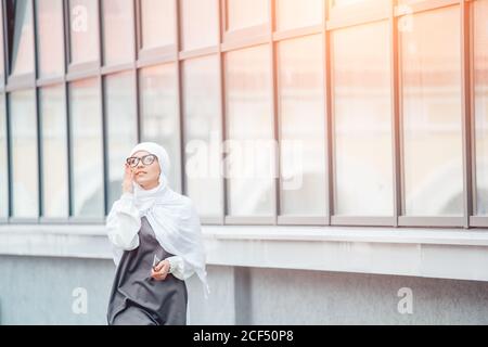 Alone muslim girl in white hijab, with glasses walking and looking in the sky. near building. outdoor Stock Photo