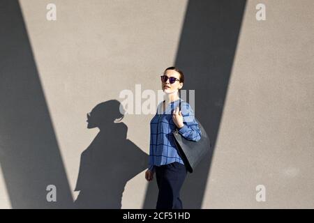 Full body female manager in sunglasses and elegant outfit smiling and looking at camera while standing against gray building wall on city street Stock Photo