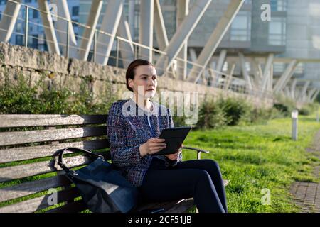 Serious female entrepreneur in elegant jacket frowning and using tablet while sitting on bench on sunny day in city park Stock Photo