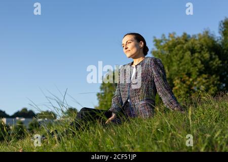 businessWoman in elegant jacket touching neck and looking away while sitting on grass against bushes in park and relaxing Stock Photo