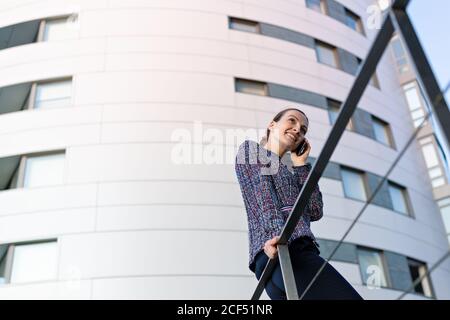 Cheerful female entrepreneur smiling and looking away while talking on smartphone and standing on background of modern buildings and cloudless blue sky Stock Photo