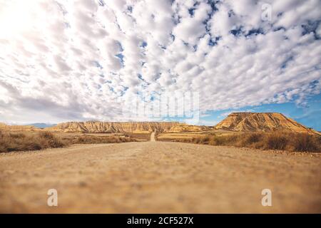 Empty road with brown soil goes into distance among dry shrubs and brown mountains and blue sky with white clouds on background at Bardenas Reales Stock Photo