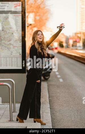 Side view of beautiful young female in trendy outfit smiling and waving hand while trying to stop taxi on city street Stock Photo