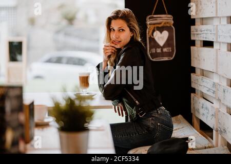 Side view of long haired trendy beautiful blonde Woman sitting in a cafe shop drinking from a glass of delicious foamy coffee Stock Photo