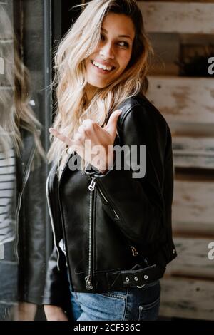 Trendy long haired cheerful Woman in leather jacket and cap leaning on table and looking away Stock Photo