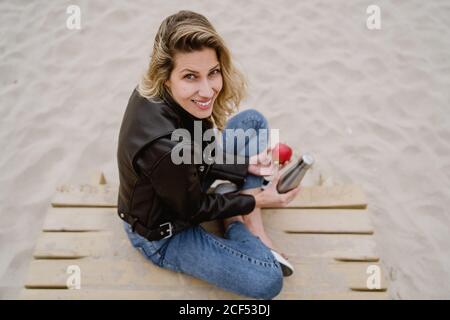 From above trendy blonde Woman in leather jacket eating red ripe apple on sandy beach looking at camera Stock Photo
