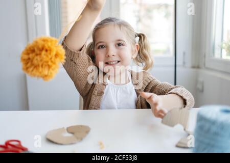 Happy little girl in warm sweater looking at pompon after finishing creating handmade craft from orange woolen threads in modern studio Stock Photo