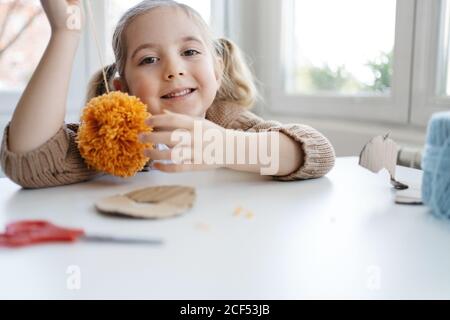 Happy little girl in warm sweater looking at pompon after finishing creating handmade craft from orange woolen threads in modern studio Stock Photo