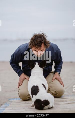 Adult male in warm jacket embracing spotted French Bulldog while sitting on wooden pier and admiring view of rippling sea on dull day Stock Photo