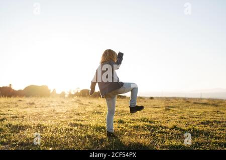 Back view of anonymous adorable active little girl in casual wear playing and dancing in green field while enjoying sunny summer evening in countryside Stock Photo