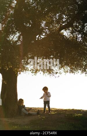 Side view of little girl with ukulele sitting with brother under big tree in field while resting together and talking in summer day Stock Photo