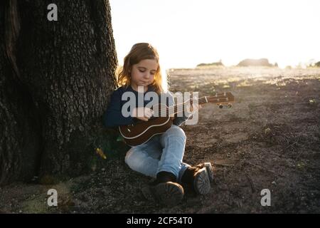 Adorable serious little girl in casual wear playing ukulele guitar while sitting near tree in sunny summer day in countryside Stock Photo