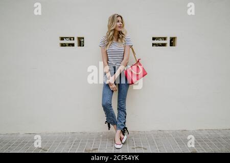 Charming young long haired blonde female with red shoulder bag wearing striped shirt and jeans standing with folded hands and looking away with shyly smile Stock Photo