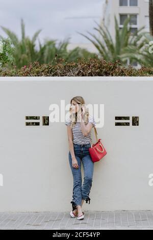 Charming young long haired blonde female with red shoulder bag wearing striped shirt and jeans standing and looking away Stock Photo