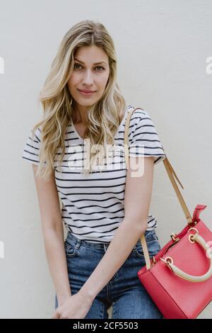 Charming young long haired blonde female with red shoulder bag wearing striped shirt and jeans standing with folded hands and looking at camera Stock Photo