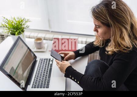 From above of focused female specialist in casual wear working with electronic documents on laptop while sitting at table near window in modern room at home Stock Photo