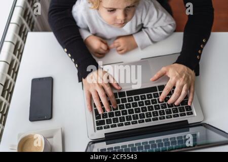 From above of crop anonymous female remote employee holding curious little child on knees while sitting at table and working with laptop at home Stock Photo