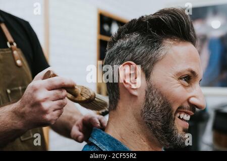 Side view of crop barber with brush near happy handsome stylish male sitting in chair in salon Stock Photo