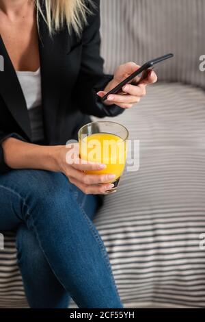 Cropped unrecognizable blonde Woman in casual clothes enjoying fresh orange juice and browsing smartphone while sitting on couch at home Stock Photo
