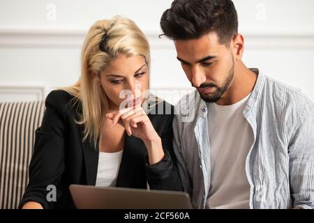 Blonde Woman and ethnic bearded handsome man in casual clothes sitting on sofa and browsing laptop together at home Stock Photo