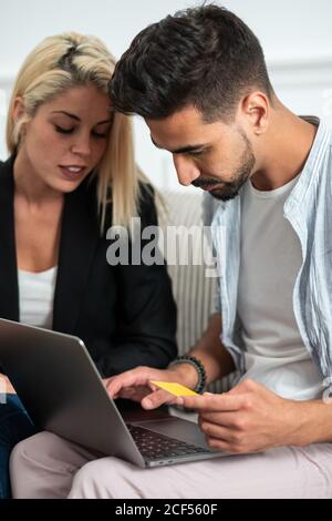 Young multiethnic couple sitting on sofa and making online purchases together with credit card on a laptop Stock Photo