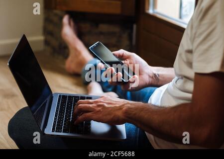unrecognizable man sitting on floor and using laptop and smartphone while working on remote project at home Stock Photo