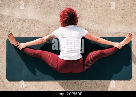 From above top view back view of anonymous barefooted Woman in sportswear doing yoga in wide angle seated forward bend pose on mat training alone on street Stock Photo