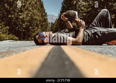 Side view of positive bearded man in sunglasses smiling and lying on back on concrete lane surrounded with leafy trees in USA Stock Photo