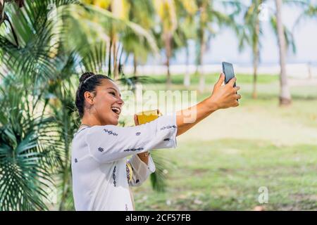Happy Woman taking a selfie with smartphone while holding mug and standing on sunny lawn by palm tree in Costa Rica Stock Photo