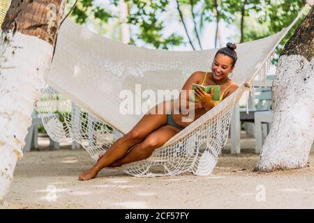 Delighted Woman in swimsuit sitting using mobile phone in hammock at exotic seaside in Costa Rica Stock Photo