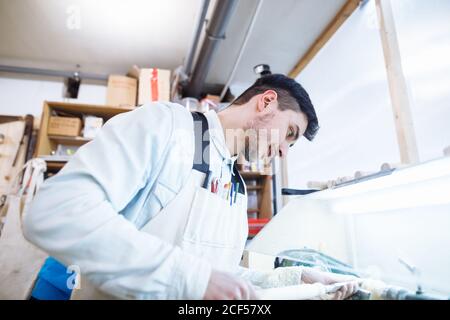 young man with dark hair and goggles holds a stalker in his hands and processes a wooden product on a lathe in the workshop Stock Photo