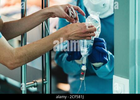 anonymous crop medical nurse giving infusion system to doctor in protective costume and mask in modern clinic during work Stock Photo