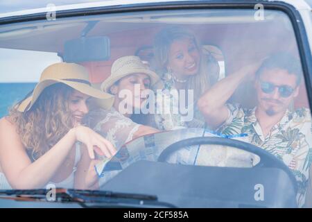 Lively cheerful ladies in hat pointing on road in map while man trying to choose right way in car in bright day Stock Photo