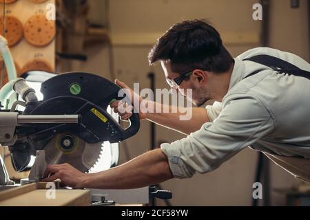 Young brunette man in black overalls by profession carpenter builder saws with a circular saw a wooden board on a wooden table in the workshop Stock Photo