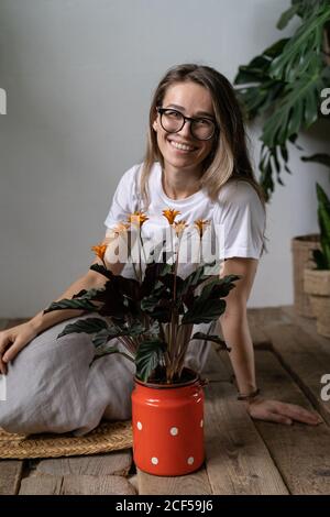 Happy woman gardener in eyeglasses wearing linen dress, sitting on the wooden floor near flowering calathea plant in old red milk can. Home garden Stock Photo