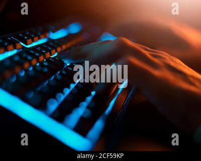 High angle of unrecognizable man in casual clothes typing on black keyboard with comfortable blue back light while using computer in dark room with dim light at night Stock Photo
