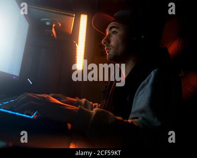 Side view of focused male in cap and wireless headset wearing casual outfit sitting alone at computer and playing video game in dark room with dim blue light at night Stock Photo