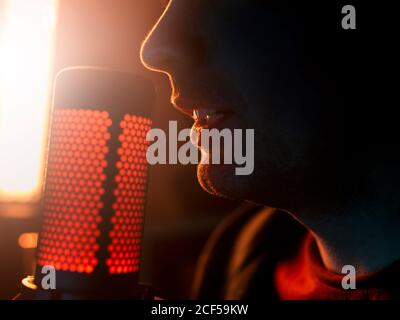 Side view cropped unrecognizable millennial male artist performing song alone beside professional mic against bright yellow longed lamp in dark studio Stock Photo